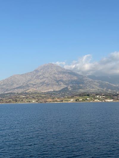 The image displays a coastal landscape in Kamariotissa, Greece. The foreground consists of a calm body of deep blue water, showing horizontal ripples across its surface. Directly behind the water, a strip of land extends horizontally across the frame. This land features a mix of green and brown vegetation, with approximately twelve small, dispersed white and light-colored buildings situated along the immediate shoreline. Behind the coastal strip, a large, imposing mountain range dominates the background. The mountain slopes are primarily grey and rocky, interspersed with patches of green vegetation, particularly on the lower elevations. The peak of the main mountain is rugged and exhibits distinct rock formations. White and light-grey clouds are present around the upper right slopes of the mountain. The sky occupies the upper portion of the image, appearing clear and bright blue with minimal cloud cover in the upper left, transitioning to more distinct cloud formations towards the right. No individuals, vehicles, or specific actions are discernible within the scene.