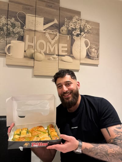 An adult male is depicted indoors in Lingolsheim, France, presenting an open box of baklava. The man has short, dark, curly hair and a full beard, with a smiling expression. He wears a black short-sleeved t-shirt featuring "CORE™ DEPT." text, a silver-toned watch on his left wrist, and a large, intricate tattoo covering his left forearm and bicep, which includes a scales-of-justice symbol and what appears to be a mythological creature. He holds a white cardboard box, labeled "Dilim" in red, filled with various pieces of baklava, characterized by golden-brown pastry, visible syrup, and green crushed pistachios. The background features a five-panel wall art piece with a sepia-toned, rustic theme, depicting items such as a watering can labeled "FLOWERS & GARDEN," a mug, a box with "HOME" inscribed, a wicker heart, green apples, candles, a pitcher of daisies, and a wicker candle holder. The wall is light-colored.