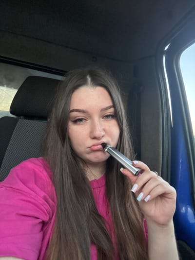 A young woman with long, dark brown hair is sitting in the driver's seat of a vehicle, likely a truck, and taking a selfie. She is wearing a bright pink t-shirt. In her right hand, she holds a silver vape pen to her lips, with her mouth pursed. Her fingernails are painted in a pastel marbled design of pink and blue. She is wearing a silver ring on her left ring finger. The interior of the vehicle is visible, including a dark gray headrest and seat with a textured pattern. The blue interior of the vehicle's door is also visible to the right of the woman. The background shows a bright, hazy sky, suggesting it is daytime. The scene appears to be in Krasnokamsk, Russia.