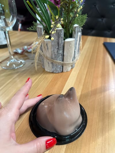 On a wooden table, a woman's hand with red nail polish holds a black plastic container with a brown, dome-shaped dessert inside. A wooden decorative centerpiece with artificial flowers and a wine glass are also visible. The location is not specified but could be in Peresvet, Russia.