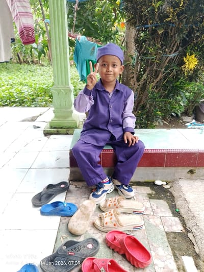 A young boy wearing a purple hat and matching shirt and pants sits on a ledge, holding up a peace sign. He wears white and blue sneakers. A collection of flip-flops and sandals in various colors are scattered on the ground in front of him. The boy is near a green pillar, and lush greenery is visible in the background. A date and time stamp is present on the bottom of the image.