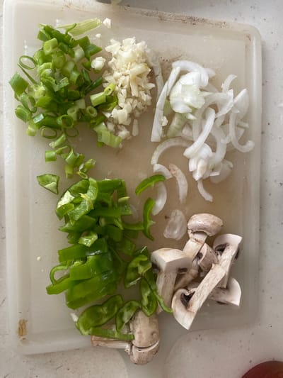 The image shows a white cutting board with various chopped vegetables arranged in separate piles. The vegetables include sliced white onions, chopped garlic, chopped green onions, sliced green bell peppers, and sliced mushrooms. The scene is likely indoors, with the cutting board placed on a countertop. The lighting suggests it could be daytime. There are no people visible in the image. The overall impression is one of food preparation, likely for a meal. The location context is Başakşehir, Turkey, but there are no specific visual cues in the image to confirm this.
