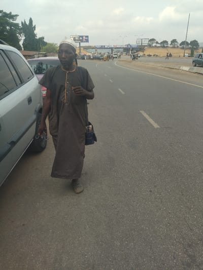 The image shows a man wearing a brown robe and a white cap walking on a paved road. He is carrying a blue bag over his shoulder. The road is empty except for the man and a car in the background. It is a cloudy day and the sky is grey. The road is lined with trees and buildings in the distance. The scene is taken in Maraba, Nigeria, located at latitude 9.024 and longitude 7.573.