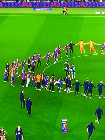 The image depicts a top-down view of a soccer field, presumably in Sant Martí Sarroca, Spain, given the context. Two teams of soccer players are visible, one team wearing blue and purple jerseys, and the other in a combination of white, green, and black. The players appear to be in a line or formation, holding hands. A few individuals in blue suits are also present on the field. There's also a flag of FC Barcelona.