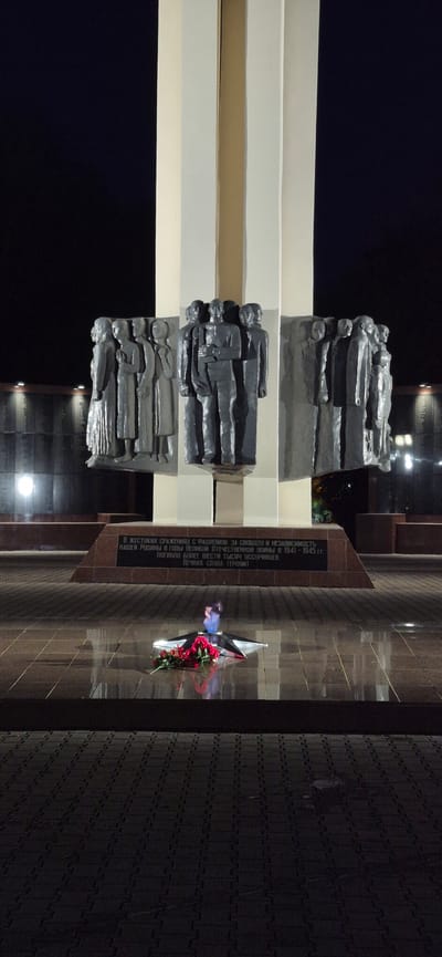 This media file depicts a monument at night, likely a memorial dedicated to soldiers. The monument features several tiers of sculptures, each depicting a group of figures lying down, possibly representing fallen soldiers. The sculptures are dark and appear to be made of metal or stone.
The scene is illuminated by artificial lighting, casting shadows and highlighting the textures of the monument and its surroundings. To the right of the monument, a wall is visible, adorned with a star-shaped emblem and a bouquet of red flowers, suggesting a memorial for a military event.
There is visible text inscribed on a dark plaque on the wall. The text is in Russian and appears to be a dedication, mentioning "cruel battles with fascism," "our Motherland," "the Great Patriotic War," and "more than six thousand Ussuriysk residents" who perished. It also includes the phrase "Eternal glory to the heroes!"
The overall atmosphere is somber and commemorative, appropriate for a memorial site. The time of day is clearly nighttime, and the setting appears to be an outdoor public space. The mention of "Ussuriysk" in the text indicates the location is Ussurijsk, Russia. The weather cannot be determined from the image.