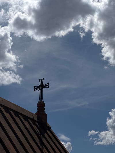 A low-angle shot of the Holy Mother of God Church in Arinj, Armenia shows a black cross affixed to the church roof, set against a cloudy blue sky. The roof is constructed with ribbed, dark-colored material. Light is inconsistent throughout the image.