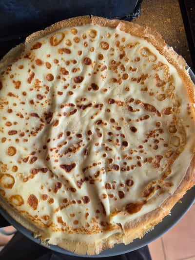An overhead, close-up photograph displays a stack of at least three circular, thin pancakes or crepes resting on a dark grey plate. The topmost crepe is predominantly a pale cream color, marked with numerous irregular, medium to dark brown caramelized spots across its surface. The texture appears slightly undulating with fine wrinkles. The outer edge of the top crepe is crisp, thin, and displays a more uniform golden-brown hue, with some areas exhibiting a slightly ruffled or torn appearance. The dark grey plate is partially visible along the lower left and right perimeters of the stacked food. In the background, to the upper left, a dark, matte, and subtly textured surface is present. To the upper right, a dark, somewhat reflective surface with visible texture, consistent with a stovetop or pan, is depicted. The lower right background reveals a section of light brown tiled flooring, while the lower left shows a glimpse of dark, soft fabric. No individuals, specific actions, or interactions are visible. The image contains no identifiable information regarding its geographical location, including the city of West Suffolk, United Kingdom.