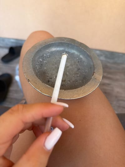 A close-up shot shows a hand with white-painted fingernails holding a cigarette. The cigarette is placed in a small, circular metal ashtray. The ashtray contains ash and cigarette butts. The background is blurred, but suggests an outdoor setting with what appears to be a paved surface and some footwear. The lighting is natural and bright. The scene is consistent with an outdoor smoking area, potentially in a residential or public space within Novi Iskar, Bulgaria.