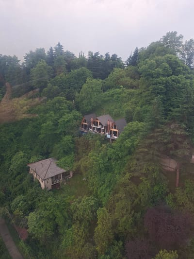 The image shows a high-angle view of a densely forested hillside with two distinct residential structures in the city of Kvreda Sameba, Georgia. The dominant element of the scene is the lush green foliage, characteristic of a temperate climate, with a variety of trees and dense undergrowth. The sky is overcast and appears hazy, suggesting either a cloudy day or fog.
The first structure, a cluster of three modern houses with dark, sloped roofs and prominent wooden beams, is situated higher up on the hill. These houses appear to be well-maintained with balconies and outdoor seating areas visible.
Further down the slope, to the left of the cluster of three houses, is a second, older-looking house. This structure has a gray, tiled roof, beige walls, and appears somewhat neglected, with vegetation encroaching on its perimeter. It has a distinct porch and a garage door.
A paved road or pathway is visible on the far left of the image, running along the edge of the property. The overall impression is one of secluded, nature-integrated living within a hilly, verdant environment. There are no people visible in the image.