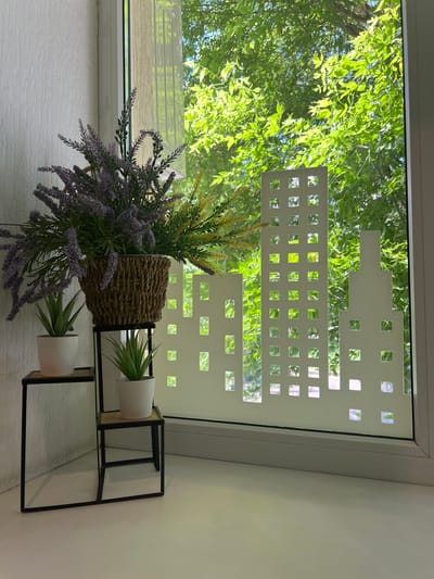 A close-up shot shows a window with a decorative film applied to the glass, depicting a white silhouette of a city skyline. In front of the window, a tiered black metal plant stand holds three potted plants. The top shelf of the stand has a large woven basket filled with artificial lavender and green foliage. Below it, on two separate shelves, sit two small white pots containing artificial succulents. The background visible through the window is a lush green canopy of trees bathed in sunlight. The location is identified as Mineralnye Vody, Russia.