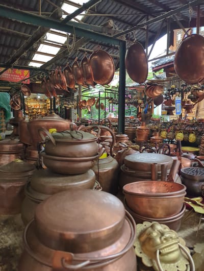 The image depicts an outdoor market stall in Gucheng District, China, filled with a vast assortment of copper cookware and decorative items. The stall is covered by a corrugated metal roof supported by green metal beams, with translucent panels allowing natural light to filter through. Rows of large, oval-shaped copper pots hang from the ceiling, along with smaller bowls and ladles. In the foreground, stacks of various-sized copper pots and kettles are displayed. One prominent object in the lower right corner is a decorative brass lion head door knocker. The background reveals more shelves filled with copper items and a glimpse of traditional Chinese architecture, including tiled roofs. The overall impression is one of a traditional marketplace showcasing artisanal copper goods. There are no people visible in the frame.
