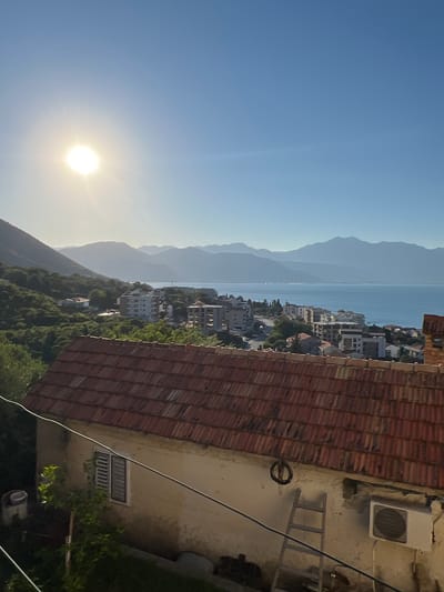 The image shows a view overlooking a coastal town in Đenovići, Montenegro, with the sun low in the sky and casting a warm glow. In the foreground, a rustic building with a red-tiled roof dominates the lower portion of the frame. A ladder leans against the wall of this building, next to an air conditioning unit. A window with slatted shutters is visible.
Beyond the foreground building, the scene opens up to reveal a town with several multi-story buildings, some appearing to be hotels or apartment complexes, interspersed with trees and vegetation. A road winds through the town. Further in the distance, a body of water, likely the Adriatic Sea, stretches out, reflecting the blue sky. Behind the water, a range of mountains forms a hazy, layered backdrop, with the sun illuminating the peaks from the left. The overall atmosphere is one of a serene, sun-drenched coastal evening.