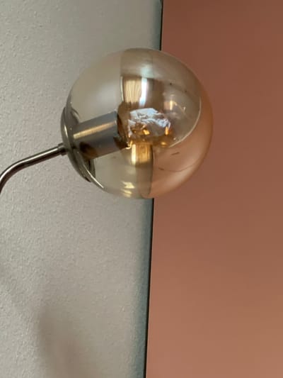 A close-up, low-angle, interior shot shows a vintage-style Edison light bulb with a transparent, amber-tinted glass globe. The bulb is suspended from a thin, metallic arm that extends from the left side of the frame. The light bulb's filament is visible, emitting a warm, golden glow. The background is divided vertically into two distinct sections: on the left, a textured, light gray wall, and on the right, a smooth, muted rose-colored wall. The scene appears to be indoors, possibly in a home or commercial space. The image does not contain any discernible elements that would place it specifically in Zelenogradsk, Russia.