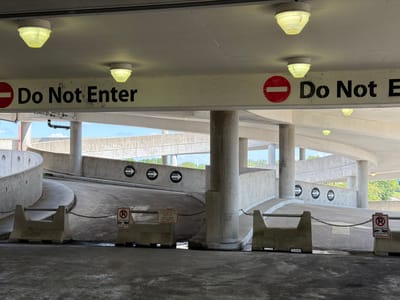 This media file captures a view of an outdoor parking garage ramp in Charlotte, North Carolina. The scene is dominated by concrete structures, including the ramp itself, large support columns, and protective barriers. Above the ramp, a concrete beam displays prominent "Do Not Enter" signs, each accompanied by a red circle with a white horizontal bar. These signs are illuminated by overhead lights.

Along the curving wall of the ramp, multiple "One Way" signs with black arrows are visible. Smaller signs indicating "No Parking" with a tow truck symbol are also present, chained between concrete barriers. One barrier has a more detailed sign with text providing a towing company's name, address, and phone number.

The time of day appears to be daytime, as natural light streams into the garage, highlighting the textures of the concrete and the shadows cast by the structures. The sky visible in the background is bright, suggesting fair weather. The overall impression is one of a functional, perhaps somewhat dated, parking facility. There are no people visible in the image.