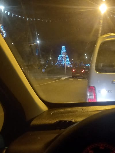 A view from inside a car, through the windshield at night. A streetlight shines brightly to the left, illuminating a decorated tower structure further away, possibly for the holidays. Cars are visible outside the windshield, with a white car directly ahead, its taillight glowing red. A dashboard with vents is visible in the foreground to the right of the shot. There is decorative bunting hanging from the upper corner of the windshield.