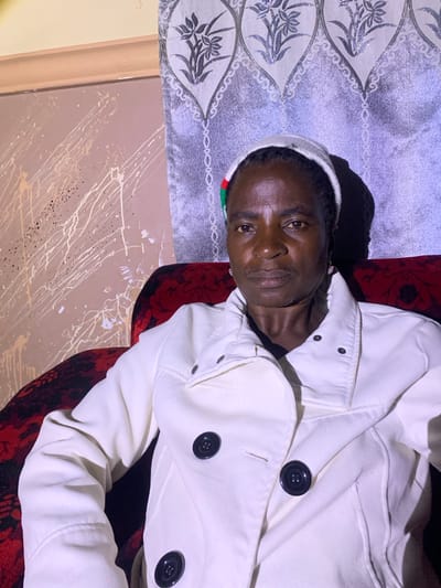 A middle-aged Black woman with dark skin and short, dark hair is sitting in a red armchair. She is wearing a white coat with black buttons and a white cap with a red, green, and black stripe on the front. She is looking directly at the camera with a neutral expression. Behind her, there is a wall with a light brown, textured paint finish with white and cream splatters. To the right of the wall is a gray curtain with a floral pattern. The lighting suggests an indoor setting, possibly a home, in Barakin Sabawa, Nigeria.
