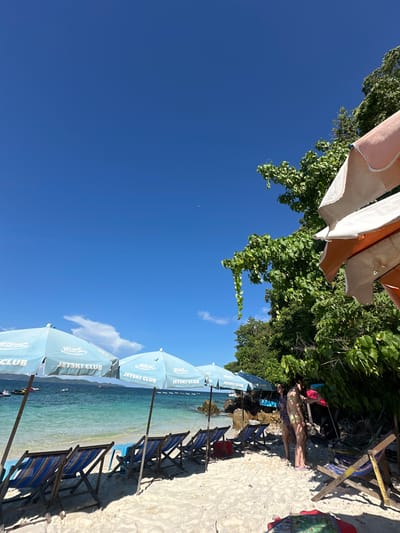The image depicts a tropical beach scene under a clear blue sky in Phru Nai, Thailand. The foreground is a sandy beach with several empty lounge chairs set up, some with blue and striped cushions. A row of light blue beach umbrellas, each emblazoned with "JETSKI CLUB," are positioned behind the chairs. The water is a vibrant turquoise, with small waves lapping at the shore. In the middle ground, several jet skis are visible in the water, and a boat can be seen further in the distance. Lush green trees and foliage overhang the beach from the right side of the frame, providing shade. Two women are present on the beach, standing near the trees and in conversation. One woman is wearing a bikini, and the other is wearing a patterned dress. The overall atmosphere is one of a sunny, recreational beach destination.