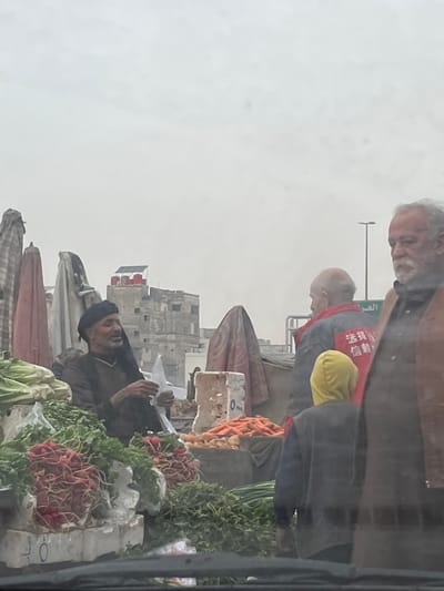 The image displays an outdoor market scene under an overcast sky. The overall lighting is soft and diffused, suggesting a cloudy day. The image appears to have been taken from a slightly elevated vantage point, possibly from inside a vehicle, with a faint reflection visible on the far right edge.

In the foreground and midground, three men are visible amidst market stalls laden with fresh produce.
On the upper left, an older man with grey hair and a beard is seen in profile, looking downwards and slightly to the right. He is wearing a dark top.
To the right of him, another man, balding or with very short hair, is also looking down towards the market stall. He is wearing a dark jacket, and a bright yellow hood or cap is visible peeking from under his collar. Most notably, he wears a red bib or sash across his chest, on which white Chinese characters are printed. The visible characters are "法治社会" (Fǎzhì Shèhuì), which translates to "Rule of Law Society" or "Law-governed Society."
Further down and towards the center-right of the image, a third man with a dark cap or head covering and a beard is looking upwards and to the left. He appears to be holding something white, possibly a plastic bag or paper, with both hands. He is positioned behind a display of vegetables.

The market stalls are filled with various fresh vegetables. Prominently displayed are piles of bright orange carrots and numerous bunches of red radishes, many with their green tops still attached. Other leafy green vegetables, possibly lettuce or other greens, are also visible. The produce is arranged in white styrofoam or plastic crates, some of which have numbers like "0" written on them.
In the lower part of the image, folded or partially open market umbrellas or awnings are visible, providing shade or protection for the stalls.

In the background, behind the market activity, several multi-story buildings are visible. They appear to be older structures, adding to the urban or semi-urban setting of the scene. On the rooftop of one of these buildings, a solar panel or water heater system can be faintly discerned.

The scene captures a mundane but vibrant moment of daily commerce in what appears to be a street market.