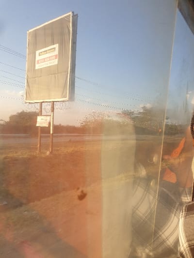 A billboard stands tall against a clear, blue sky. It displays a faded advertisement with the words "Tellow Records" and a phone number visible. Behind the sign, a road stretches into the distance. A barbwire fence runs alongside the billboard. The picture is taken through a dirty window, obscuring the view and adding a vintage, almost distorted effect. A person wearing a plaid shirt is visible in the reflection.