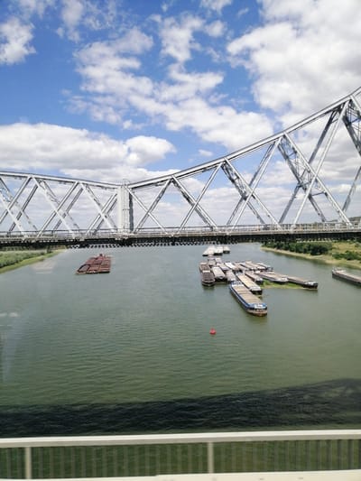 This is an aerial, eye-level, medium shot of the Danube River in Cernavoda, Romania, on a partly cloudy day. A large, modern steel truss bridge spans the river horizontally across the middle of the frame, with its intricate metalwork dominating the upper portion. Below the bridge, the river is calm and green, reflecting the sky. Several barges are moored in the river, some clustered together and others more spread out. On the left side of the frame, a long convoy of barges is tied up. On the right side, a single barge is positioned further away, and another longer barge is visible in the far distance. Green, tree-covered banks line the river on the far side. The foreground of the image shows a white railing, suggesting the photo was taken from another bridge or a high vantage point. The sky is a vibrant blue, dotted with fluffy white clouds, some of which cast faint shadows on the water. There are no people visible in the scene.