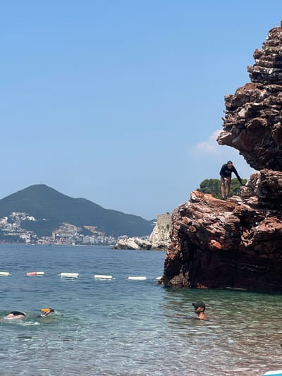 The image captures a dynamic scene at Pržno, Montenegro, on a clear, sunny day. The foreground is dominated by a rocky cliff face, with a person poised to jump into the turquoise water below. This individual is wearing a black long-sleeved shirt and patterned shorts, demonstrating an active pursuit of adventure.
In the water, another person is visible, wearing a dark cap and appearing to swim or float. Further out, a person is wearing snorkel gear, with an orange snorkel. The water is calm and inviting, with a series of white and orange buoys marking a section of the sea.
Behind the immediate cliff and water, a densely built-up town is nestled against a verdant mountain slope. The architecture suggests a Mediterranean coastal settlement. The overall impression is one of a vibrant summer day at a popular coastal destination, with opportunities for both cliff jumping and water activities. The clear blue sky indicates pleasant weather.