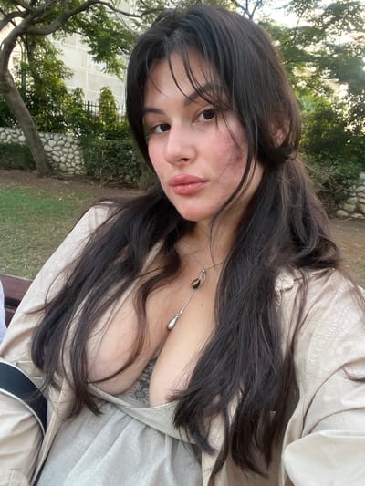 A young woman with long, dark hair is captured in a selfie-style shot. She is outdoors, with a blurred background of greenery, trees, and a stone wall, suggestive of a park or garden setting. The sun appears to be casting dappled light, indicating it's daytime.
The woman is wearing a light-colored, possibly linen, outfit that is open, revealing a portion of her chest. A delicate necklace with a teardrop-shaped pendant hangs around her neck. She has a soft expression, with her lips slightly parted and her gaze directed towards the camera. Her makeup appears natural, with emphasis on her lips and cheeks. A subtle tattoo is visible on her upper chest.
The scene is set in Kiryat Motzkin, Israel, suggesting an outdoor leisure activity. The overall mood is relaxed and informal. No text is visible in the image.