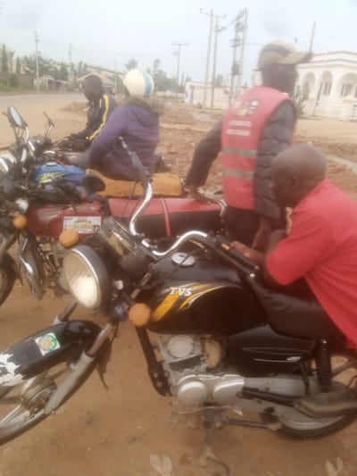 In Television, Nigeria, a group of individuals are gathered around motorcycles on a dirt road. The scene is overcast, suggesting a cloudy or hazy day. In the foreground, a person wearing a bright red shirt sits on a black motorcycle with "TVS" branding. The motorcycle has a prominent headlight and orange turn signals. Behind this motorcycle, another person in a red vest and a tan cap stands, with a blurred individual in a purple jacket and white helmet seated on a maroon motorcycle. This maroon motorcycle, labeled "BOXER," has a sticker with an image of people. The background reveals a street with utility poles and a white building with arched windows. The overall impression is a casual gathering or waiting area for motorcycle transport.