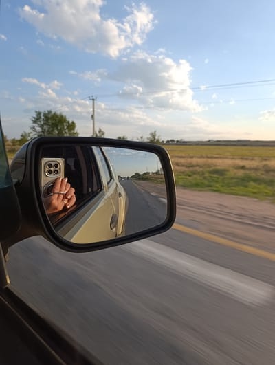 The image is a reflection in a car's side mirror, showing a person's hand holding a smartphone. The hand has a ring and the phone case has multiple camera lenses. The car is in motion on a paved road with a yellow line. The background is a rural landscape with dry grass, trees, and a power pole under a cloudy sky. The city of Rassvet, Kazakhstan is not visible in the image.