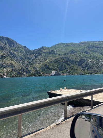 View of the Bay of Kotor from a road in Prčanj, Montenegro. A mountain covered in lush green vegetation rises from the shore on the left. The blue water of the bay occupies the center of the frame. A grey metal guardrail runs parallel to the road, which is made of concrete or asphalt. Part of a car, specifically a side mirror, is visible on the right edge of the frame, reflecting the environment and indicating that the photo was taken from inside a vehicle.