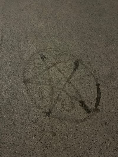 The image displays a wet, circular marking on an asphalt surface, likely a street or pavement in Zabrze, Poland. The marking appears to be a pentagram, a five-pointed star enclosed within a circle, drawn with liquid. The asphalt is dark grey and textured, indicating it is an outdoor urban environment. No people or other objects are visible in the immediate vicinity of the marking. The lighting suggests it may be nighttime or dusk.