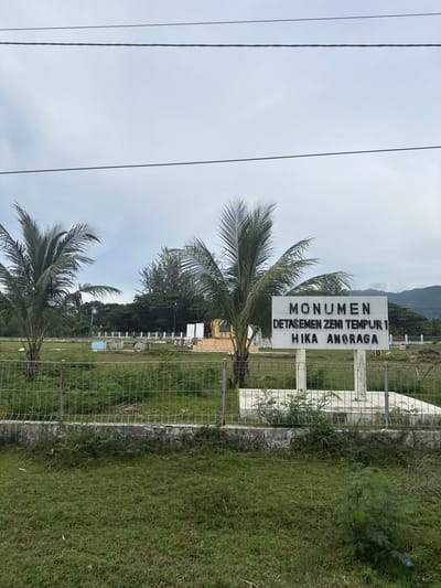 The image depicts an outdoor scene at the city of Lamkruet, Indonesia. In the foreground, a metal fence separates a grassy area from a field of green vegetation. Beyond the fence, a white monument stands with black lettering that reads "MONUMEN DETASEMEN ZENI TEMPUR 1 HIKA ANORAGA". Two palm trees are visible to the left of the monument. In the background, there are more trees and some structures, with hills in the far distance under a cloudy sky. Power lines are visible at the top of the image. The environment appears to be a rural or semi-rural area with a monument and natural vegetation.
