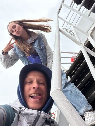 Two individuals, a man and a woman, are captured in a playful, upward-angle photograph, likely a selfie or taken from below, set against an overcast sky. The woman, with long brown hair flowing downwards, is on the left, looking directly at the camera with a slight pout and making a peace sign with her right hand. She is wearing a light-colored top under a blue denim jacket. The man is on the right, wearing a grey and navy hooded jacket, sticking out his tongue in a humorous expression.

They are positioned beneath a white metal structure, possibly part of a boat or ferry deck, which features railings and a black slotted overhead area. A glimpse of a red lifebuoy is visible in the upper right background, further suggesting a marine setting. The lighting indicates daytime, but the sky is uniformly grey, suggesting an overcast or cloudy day. No text is visible in the image. The location context provided is Nizhneleninskoe, Russia.