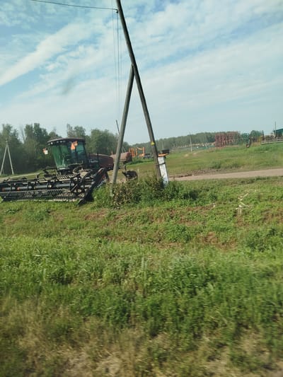 In an outdoor scene in Урожайный, Russia, a large, maroon combine harvester with a wide header attachment is parked in a grassy field. The machine, identified by "MacDon" branding on its header, is positioned to the left of center in the frame. Behind the combine, a line of trees forms the backdrop. In the middle ground, a dirt road or track is visible, and further in the distance, rows of what appear to be agricultural equipment, possibly seeders or plows, are stored. The sky above is partly cloudy with a blue expanse. The foreground is filled with green grass, with some areas of dry, brown vegetation. A utility pole with wires runs vertically through the left side of the image.