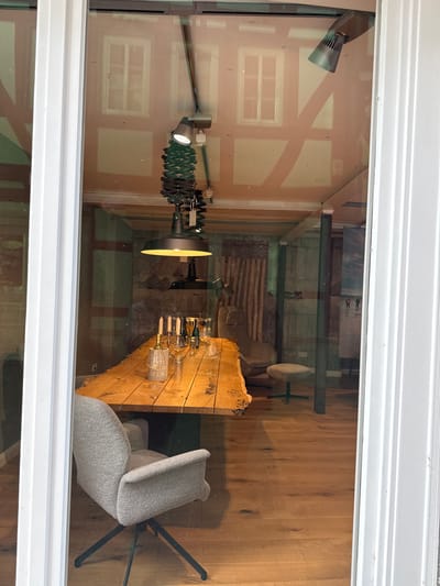 The image shows a window display of an establishment in Wetzlar, Germany. Inside, a large, rustic wooden table is set with wine glasses, bottles, and candles. A grey armchair with a dark metal base is positioned to the left of the table. Behind the table, a bamboo curtain hangs against a textured wall. A small stool with a grey cushion is visible to the right of the table. Overhead, a black industrial-style pendant lamp with a coiled green arm hangs from the ceiling. The reflection on the window shows architectural details of a building, including half-timbered beams and windows.