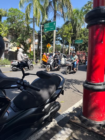 In Gianyar, Indonesia, under a clear blue sky, a street scene unfolds with numerous motorbikes and a red pole in the foreground. Several people are riding motorbikes, some wearing helmets. One person in a gray hoodie rides a silver scooter, while others wear orange and pink jackets. Palm trees line the street, and traffic signs indicate directions to "Sukawati Denpasar" and "Bone". A yellow traffic signal hangs above the road. In the background, buildings and a red truck are visible. The perspective suggests the photo is taken from another motorbike, with its handlebar and seat in the foreground.