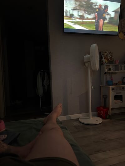 The image depicts an indoor scene, likely a living room, with a television displaying a police-related program. A person's legs are visible in the foreground, resting on a green blanket. To the right, a white floor fan stands near a child's play kitchen set and a red basket. A closet with clothes hanging inside is visible on the left. The image does not provide any information or visual cues that would allow for the determination of a specific location within the city of Syktyvkar, Russia.