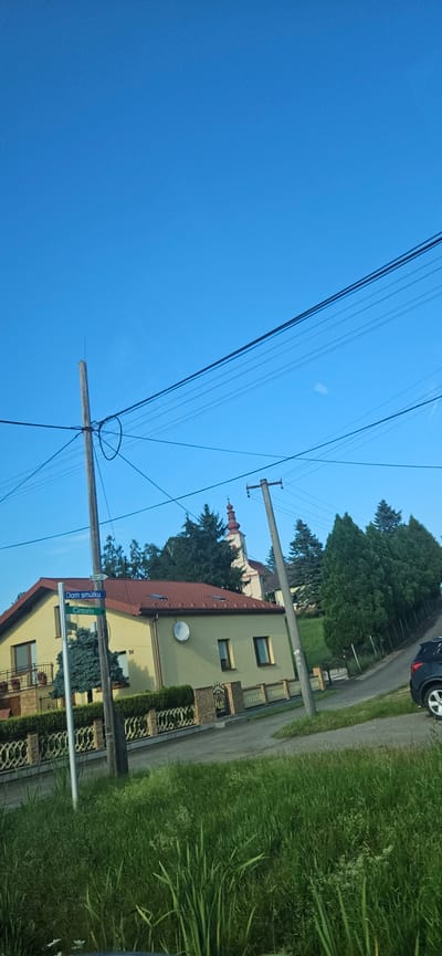 In the city of Radoma, Slovakia, on a clear blue day, a street sign reads "Dom smútku" and "Cintorín". To the right of the sign stands a house with a yellow facade and a red tile roof, bordered by a decorative stone fence. Further down the street, a church with a red steeple is visible behind a line of evergreen trees. Power lines crisscross the sky, supported by wooden and concrete poles. A dark-colored car is parked on the right side of the road, adjacent to a grassy embankment. The scene is characterized by a tranquil, suburban atmosphere under a bright sky.