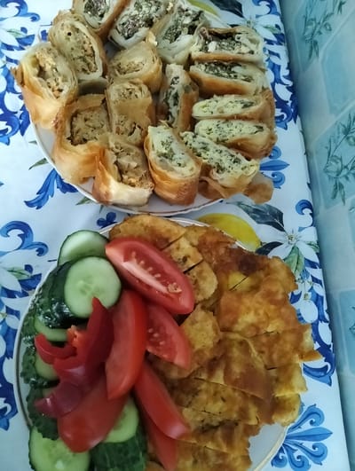 Two plates of food are presented on a tablecloth with a blue and white floral pattern. The upper plate contains several pieces of what appears to be a savory pastry, cut into bite-sized portions. The pastry is golden brown and flaky, with visible filling in each piece. The filling seems to be a mixture of finely chopped ingredients, possibly herbs, cheese, and some other elements. The lower plate holds sliced vegetables and another type of prepared food. Sliced tomatoes, red bell pepper, and cucumber slices are arranged on one side. The other side of this plate features a dish cut into pieces, which resembles a frittata or a thick omelet, golden in color and with visible herbs within. The background suggests an indoor setting, possibly a kitchen or dining area, with a tiled wall visible on the right side. The tablecloth and the presentation of the food suggest a home-cooked meal. The image captures a food spread, possibly for a meal or a gathering, at an undisclosed location in Cupcini, Moldova.