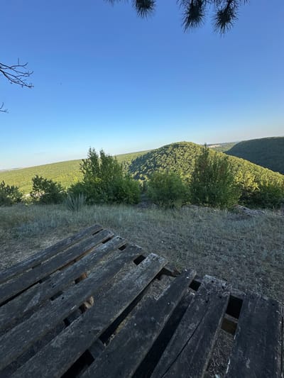 The image depicts a scenic overlook in the city of Pryvorottia Druhe, Ukraine. In the foreground, a weathered wooden structure, possibly a picnic table or bench, is positioned on dry, grassy terrain. The structure is composed of rough, dark wooden planks with visible gaps and wear. Beyond the foreground, a lush, green landscape unfolds with rolling hills densely covered in trees. The sky is a clear, bright blue with the sun casting gentle light on the scene. The overall impression is one of natural beauty and tranquility.