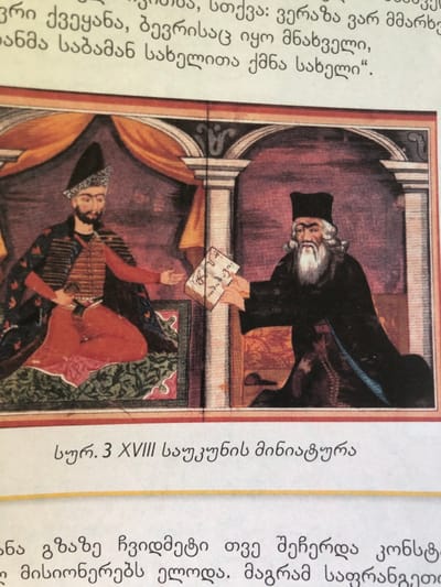 This image is an 18th-century miniature painting depicting an indoor scene set in what is contextualized as Ortabatumi, Georgia. Two men are seated, facing each other, engaged in an exchange.
The figure on the left is younger, with a dark beard and mustache, wearing a tall, dark conical hat with an ornate band. He is dressed in a dark patterned cape or robe over a striped red and black garment, and red trousers, with a dagger visible at his waist. He sits cross-legged on a green patterned rug, his right hand extended towards the other figure in a gesture of reception.
The figure on the right is older, distinguished by a long white beard, mustache, and shoulder-length white hair. He wears a dark, flat-topped hat and a long, dark robe. He is seated on a dark cushion or bench, extending both hands forward to present a rectangular object, likely a book or document, to the left figure. Georgian script is legible on the cover or pages of this object.
The background features dark red drapery, suggesting a tented interior or a richly decorated room. A fluted white column with an ornate capital stands between the two men.