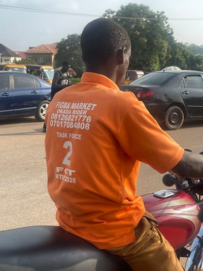 The image shows a back view of a person sitting on a red and gold motorcycle in a city street. The person is wearing an orange t-shirt with white text that reads "FIGBA MARKET OKADA RIDER 08126821776 07017084808 TASK FORCE 2 FCT MTV/2026". They are wearing brown pants. The motorcycle's fuel tank is visible, and a chrome component is on top of it. The background shows a street with several vehicles, including a blue car, a black car, and yellow tricycles, commonly known as "keke napeps" in Nigeria. Buildings and trees are also visible in the background, suggesting an urban or semi-urban setting. The sky is clear, indicating daylight. The scene appears to be in Galadimawa Village, Nigeria, based on the presence of okada riders, keke napeps, and the general streetscape.