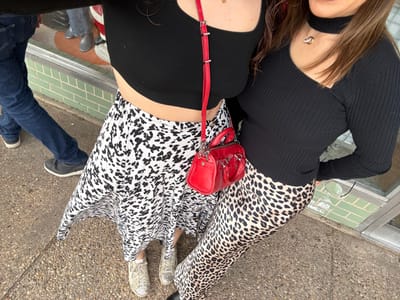 Three people are visible from the waist down, lying on a gray stone surface. One person wears brown leopard print pants and the other a black and white leopard print skirt with white sneakers. A small red purse rests on the second person's waist. Part of the third person's legs and feet are seen; they wear blue jeans and gray sneakers. The foreground contains a glass surface with green tiles.