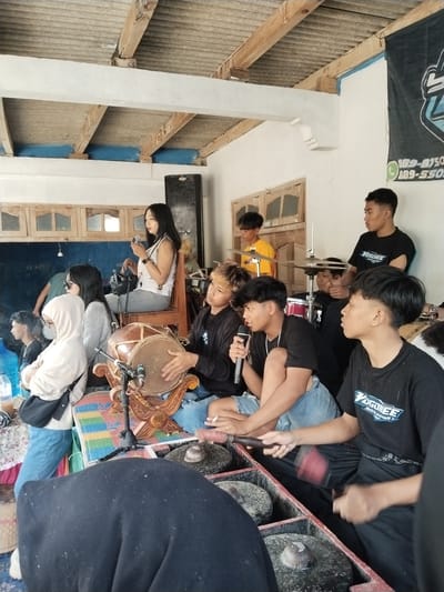 This image shows a group of people in Jenawi, Indonesia, likely during a performance or gathering. Several individuals are seated or standing around a collection of musical instruments including a kendang (drum), a set of gongs, and a drum set. Several individuals are wearing casual clothing, and one is holding a microphone. A woman sits on a wooden chair, observing the scene. A banner is visible in the background with lettering and numbers on it. The room's ceiling reveals exposed beams and a corrugated roof. Several windows and doors are visible in the background.