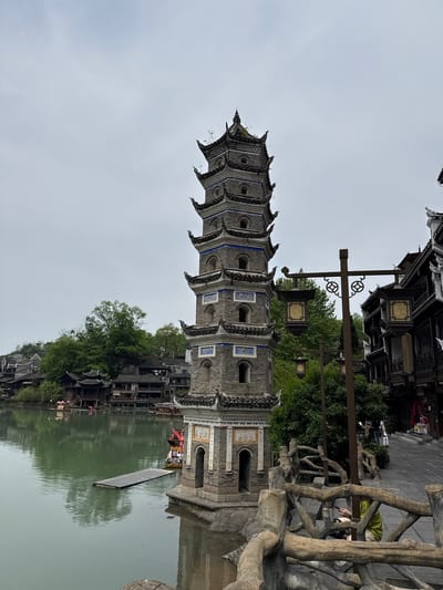 The image shows a pagoda reflected in a body of water. The pagoda is tall and narrow, with multiple tiers and a grey brick exterior. Each tier has a roof with ornate eaves. There are square openings along the upper parts of the pagoda, with a possible decorative tile pattern. The water reflects not only the pagoda but also parts of the surrounding architecture, which includes traditional-style buildings with dark roofs and walls, with trees situated between the buildings. There is a lamp post in the foreground. The sky is overcast. The overall setting is likely Fenghuang, China, known for its well-preserved ancient town and river features. A rough railing made of tree branches is on the upper right-hand side of the image. There is a boat on the lake with at least one person in it.