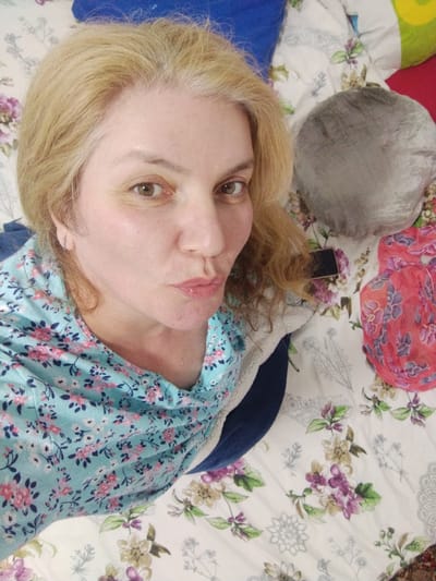 A middle-aged woman with blonde hair and light-colored eyes is taking a selfie. She is wearing a light blue top with a floral pattern and has her lips puckered. She appears to be lying down on a bed with a floral patterned comforter. There are pillows in the background, one is gray and round, another is red and a corner of a green pillow is visible. A red scarf with a floral pattern is also visible on the bed. The image is taken from a close-up, high-angle perspective. There is no information to suggest the location in Al Mas'oudi, United Arab Emirates.