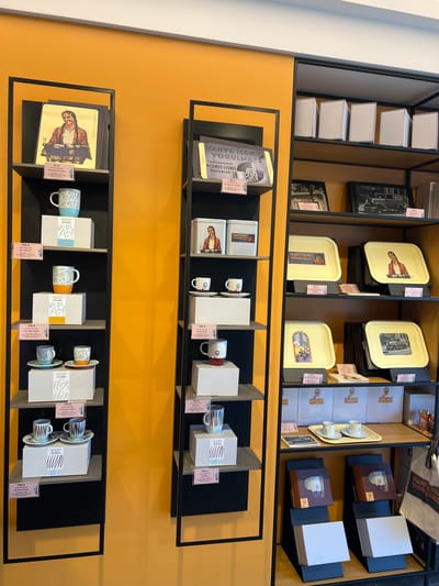 This media file captures a retail display of coffee-related items, likely in a store in Kadıköy, Turkey, given the Turkish text. The scene is set against a warm orange wall and features multiple shelves organized with products. The items on display include various types of coffee tins, boxes containing cups and saucers, and possibly books or artwork.
On the upper shelves, several rectangular tins with images are visible. One tin displays a portrait of a person in a brown jacket, while another shows a black and white photograph of a vehicle and people. Below these, more yellow tins are arranged, one featuring a stylized illustration of coffee beans and text, and another with a patterned design.
The middle shelves are dedicated to packaged goods. There are white boxes, some with dark brown sleeves, each containing what appear to be coffee cups and saucers. Some of these sets have visible text and logos, including "CE" and "KURUKAHVECİ MEHMED EFENDİ". Loose cups are also displayed, some with distinctive patterns.
The bottom shelves continue the theme with more packaged sets of cups and saucers, along with individual cups. The overall impression is a curated selection of high-quality coffee merchandise, presented in an organized and visually appealing manner. The lighting suggests an indoor setting, possibly during daylight hours, with no indication of specific weather conditions. The meticulous arrangement of the products and the clean aesthetic convey a sense of professionalism and attention to detail. The visible text is primarily in Turkish, advertising the brand "Mehmed Efendi" and phrases like "Kahve İçen Yorulmaz" (One who drinks coffee never tires).
