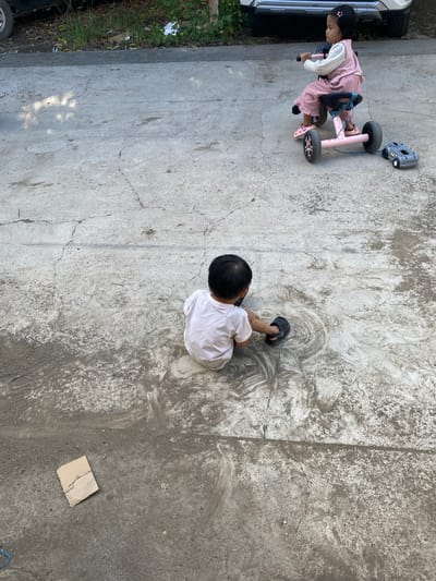 In Ta', Indonesia, two young children are present on a cracked concrete surface. The first child, a girl, is seated on a pink tricycle, wearing a pink outfit and white long-sleeved shirt. She is being towed by a gray toy car. The second child, a boy, is seated on the ground facing away from the camera, wearing a white t-shirt and black sandals. He is engaged in playing on the concrete, drawing circular patterns. The environment is outdoors, with a car and vegetation visible in the background. A torn piece of cardboard lies on the ground near the boy.