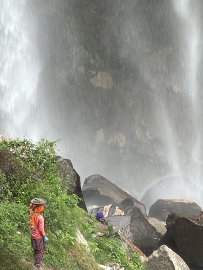 An over-the-shoulder view captures a person in a pink shirt and patterned pants, carrying a flat, round object on their head, with their back to the camera. The person stands near a lush, green embankment, beside large, dark rocks. In the background, a powerful waterfall cascades down, creating a misty, ethereal atmosphere. The scene appears to be in a natural, possibly mountainous, environment.