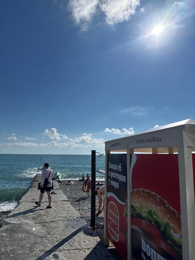 On a bright, sunny day at a beach in Adlerskiy, Russia, a concrete pier juts out into the turquoise ocean. The pier is lined with pebbles and leads to a white canopy structure advertising "Beach Chaika" and a Burger King advertisement. A man in a white shirt and black shorts walks away from the camera on the pier. Other beachgoers are visible in the background, some sunbathing on the shore. The sun is high in the sky, casting sharp shadows.