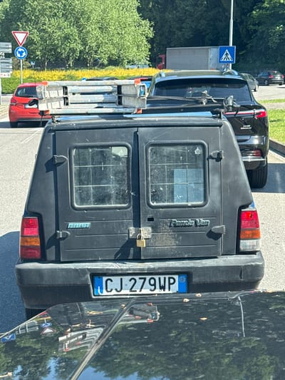 The image captures the rear view of a black Fiat Panda Van parked outdoors on a sunny day in what appears to be an urban or suburban setting in Como, Italy.
The van's rear is prominently displayed, showing its black, somewhat worn surface. The words "Panda Van" are visible on the right rear door, and "FIAT" is on the left rear door. Both doors feature small, gridded windows, suggesting security bars or a cage inside the cargo area. A golden padlock is attached to the latch on the left rear door, indicating the cargo space is secured. The rear bumper is black plastic, and the tail lights, consisting of red, orange, and clear sections, are visible on either side.
The Italian license plate, "CJ 279WP," is attached to the left side of the rear bumper. It includes the European Union symbol with the letter "I" for Italy on its left blue strip, and "WP" on a blue strip to the right.
In the background, another black car (possibly an SUV) is parked behind the van, and further down the road, a red truck with ladders secured to its roof rack is visible. Green trees and foliage line the side of the paved road.
Several road signs are present: a blue square sign indicating a pedestrian crossing, a blue circular sign for a roundabout, and a red triangular warning sign with a right-pointing arrow. The bright sunlight creates clear shadows and detailed reflections, including trees and road signs, on the shiny black car next to the van.